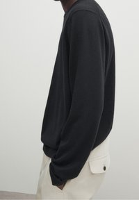 Vue de côté d'une personne portant un pull noir à manches longues et un pantalon clair avec une poche arrière boutonnée, les mains dans les poches.