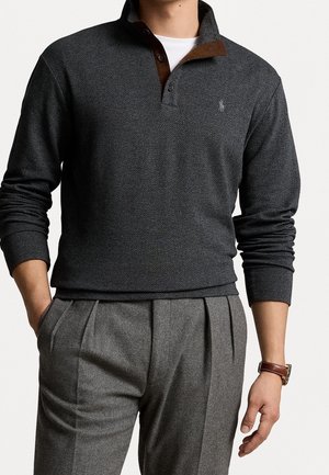 Homme portant un polo en maille chevron gris foncé sur une chemise blanche, avec un pantalon gris plissé et une montre-bracelet en cuir marron.