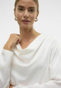 Balts blekšķis ar garām piedurknēm un kapuces apkakli, izgatavots no teksturēta auduma. Modelim ir lieli riņķi ausīs, āda ir dabiski toņos ar detaļām.