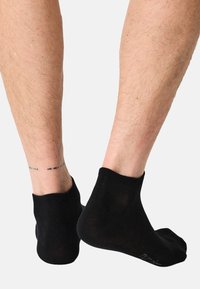 Nohy v černých kotníkových ponožkách, zobrazené zezadu a z boku, s malým černým tečkovaným tetováním na kotníku levé nohy.