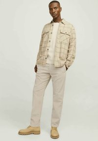 Homme debout portant une chemise à carreaux beige à boutons, un pull écru, un pantalon beige clair et des chaussures lacées beiges sur fond uni.