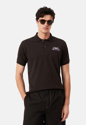 Hombre con polo oscuro con logo de Lacoste y pantalones cortos negros, posando con las manos en los bolsillos y llevando gafas de sol con montura carey.