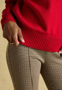 Red knitted sweater with ribbed cuffs paired with houndstooth patterned brown trousers. Close-up of hand resting on the waistband.