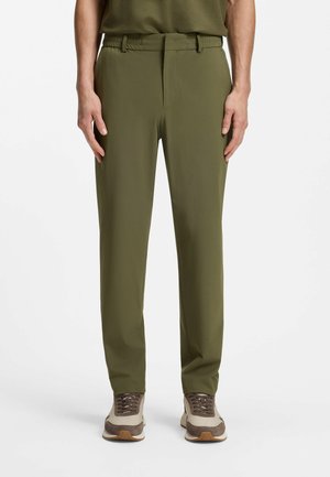 Mann trägt olivgrüne, gerade geschnittene Hose und beige Sneakers und steht vor einem schlichten weißen Hintergrund.