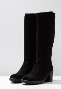 Bottes noires montantes en daim, dotées d'une forme légèrement courbée, d'une surface texturée, d'un talon épais et de surpiqûres décoratives.