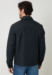 Hombre con cabello rizado oscuro, lleva una chaqueta negra y vaqueros azules, de espaldas contra un fondo claro y liso.