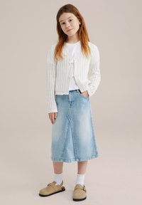 Fehér kötött kardigán nyitott mintával, világoskék denim midi szoknyával párosítva, bézs slip-on cipőkkel és fehér bokazoknival.