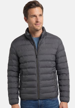 Mężczyzna w ciemnoszarej pikowanej kurtce z przednim zamkiem na błękitnym swetrze, patrzący nieco w prawo na jednolitym tle.