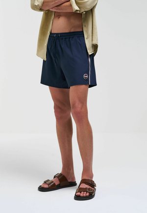 Shorts de bain bleu marine avec une taille élastique, des bandes latérales et un patch logo rond. Associés à des sandales marron à double boucle.