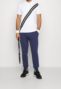 Bílá polo košile s černými diagonálními pruhy, spárovaná s tmavě modrými joggerami a černými sportovními botami. Drží tenisovou raketu.