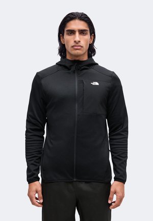 Junger Mann mit mittellangen Haaren, der einen schwarzen Kapuzenpullover mit Reißverschluss und The North Face Logo auf der Brusttasche trägt, steht vor einem schlichten Hintergrund.