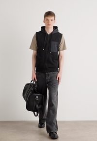 Μαύρο αμάνικο γιλέκο με γκρι τσέπη, φορεμένο πάνω από μπεζ T-shirt, σε συνδυασμό με σκούρα τζιν. Το μοντέλο κρατά μια μαύρη τσάντα χεριού.