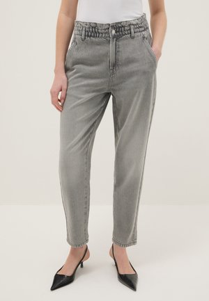 Mujer con pantalones vaqueros grises de tiro alto con cinturilla elástica, manos en los bolsillos, combinados con zapatos de tacón bajo puntiagudos negros y camiseta blanca.