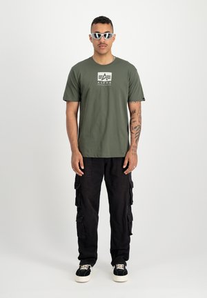 Camiseta de algodón verde con un logo blanco, combinada con pantalones cargo negros con múltiples bolsillos y zapatillas retro negras con suelas blancas.