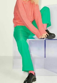 Pull côtelé rose, pantalon vert, chaussures noires et chaussettes roses. Mannequin assis sur une surface transparente, mettant en valeur un contraste de couleurs vives.
