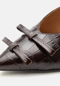 Καφέ μυτερό δερμάτινο παπούτσι με υφή και δύο διακοσμητικούς δερμάτινους φιόγκους στο επάνω μπροστινό τμήμα.