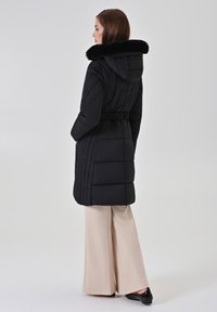 Zwarte gewatteerde donsjas met een capuchon van nepbont, een getailleerde taille en zijzakken; gedragen over lichtbeige broek met wijde pijpen en zwarte schoenen.