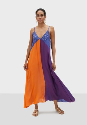 Robe maxi en violet, orange et bleu avec un décolleté en V. Présente des broderies florales sur la poitrine et une silhouette fluide. Sandales argentées visibles.