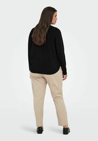 Donna con lunghi capelli castani di profilo, che indossa un maglione nero, pantaloni beige e scarpe nere con borchie su uno sfondo chiaro.