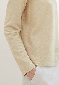 Beige långärmad topp med ett texturerat stickmönster. Mjuk, lättviktsduk som har en avslappnad passform och en fåll som slutar ovanför höfterna.