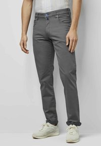 Graue Chinos mit slim Fit, versehen mit einem blauen Logo, Vordertaschen und umgeschlagenen Säumen, kombiniert mit weißen Sneakers vor einem neutralen Hintergrund.