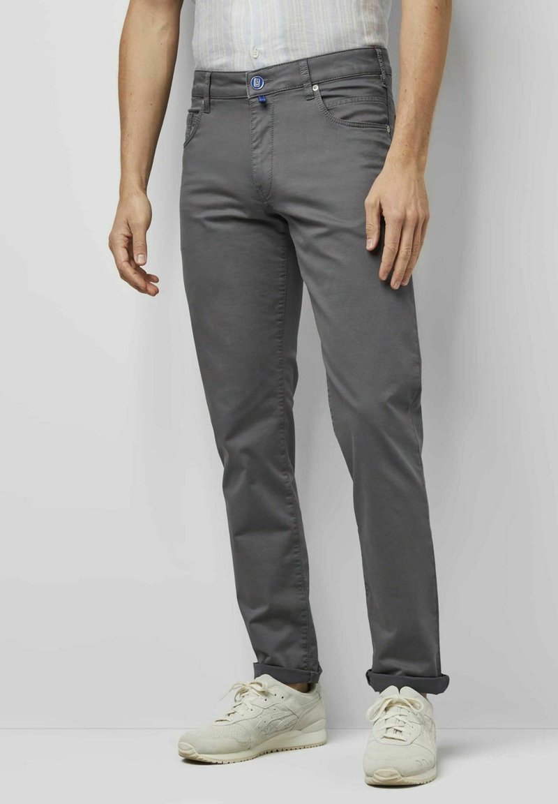 Graue Chinos mit slim Fit, versehen mit einem blauen Logo, Vordertaschen und umgeschlagenen Säumen, kombiniert mit weißen Sneakers vor einem neutralen Hintergrund.