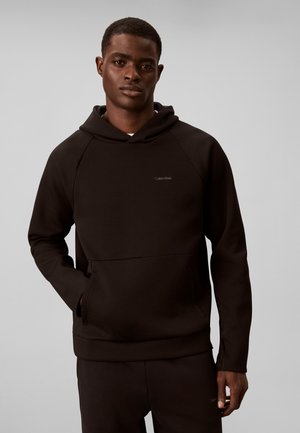 Νέος άνδρας που φοράει μαύρη φόρμα Calvin Klein με κουκούλα και μαύρο παντελόνι που ταιριάζει, στέκεται με το ένα χέρι στην τσέπη της κουκούλας.