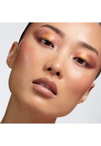 Nahaufnahme eines Gesichts mit glatter, hellbrauner Haut; warmem Rosé- und Gold-Augen-Make-up; vollen Lippen mit einem sanften Rosa; definierten Augenbrauen.