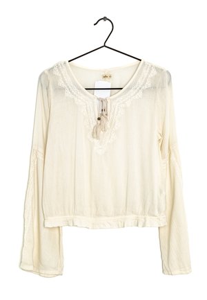 Cremefarbene Langarmbluse mit Spitzenbesatz und Quastenband am Ausschnitt, hängend an einem schwarzen Kleiderbügel vor weißem Hintergrund.