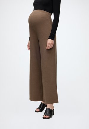 Personne enceinte portant un pantalon de maternité marron à jambes larges, un haut noir à manches longues et des sandales à talons noires à bout ouvert, debout sur un fond uni.