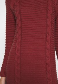 Robe en tricot bordeaux à côtes avec un motif de tricot en câble vertical sur les côtés et des manches texturées. Tissu doux et chaud.