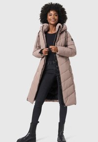 Long, padded coat in light beige with a hood, featuring a zip front, black accents, and a quilted pattern. Worn over black jeans and boots.