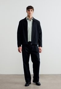 Νέος άνδρας όρθιος, φορώντας ένα ελαφρύ πουκάμισο με κουμπιά κάτω από ένα μαύρο μπουφάν με φερμουάρ, μαύρο παντελόνι και μαύρα παπούτσια, μπροστά σε απλό φόντο.