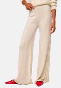 Pantalons crème à jambes larges en tissu doux, avec des détails de tresse sur les côtés et une taille haute, associés à des chaussures pointues rouges.