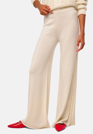 Pantalons crème à jambes larges en tissu doux, avec des détails de tresse sur les côtés et une taille haute, associés à des chaussures pointues rouges.