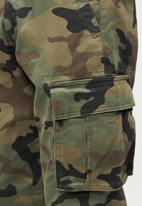 Πράσινο και καφέ καμουφλάζ παντελόνι εργασίας με τσέπη με καπάκι στο μηρό και εμφανείς λεπτομέρειες ραφής.