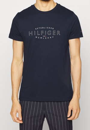 Mężczyzna ubrany w granatowy T-shirt z napisem „Established Hilfiger MCMLXXXV” i granatowe spodnie w prążki, stojący z rozluźnionymi ramionami.