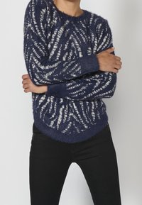Persona con los brazos cruzados, vistiendo un suéter de lana azul marino con patrones de zigzag blancos y pantalones negros, frente a un fondo liso.