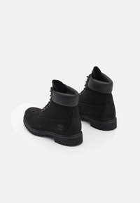 Botas de tobillo de ante negro con un collar acolchado, punta redonda y cierre con cordones. Presentan una suela de goma texturizada y un detalle de logo en el costado.