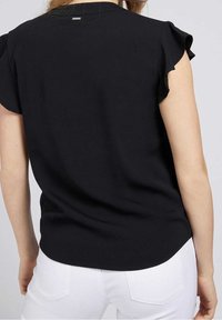 Femme portant un chemisier noir à manches courtes avec des manches volantées et un pantalon blanc, vue de dos sur un fond uni.