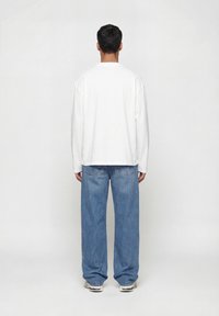 Άτομο με την πλάτη, φοράει απλό λευκό μακρυμάνικο πουκάμισο, φαρδιά μπλε τζιν και λευκά αθλητικά παπούτσια, σε φωτεινό απλό φόντο.