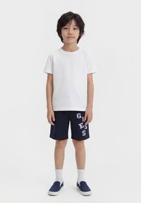 Αγόρι που στέκεται σκυφτό προς τα εμπρός φορώντας λευκό μπλουζάκι, σκούρα μπλε σορτς με γράμματα "GUESS", λευκές κάλτσες και σκούρα μπλε παπούτσια χωρίς κορδόνια.