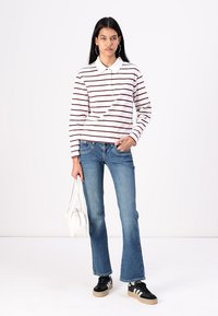 White long-sleeve polo with maroon stripes, paired with flared blue denim jeans and black sneakers. Model holds a white bag.