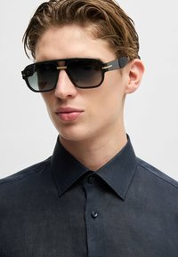 Occhiali da sole neri rettangolari con lenti sfumate, montatura accentuata in oro e aste con texture. Indossati con una camicia button-up scura.