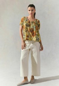 Blusa floral de manga corta en amarillo y verde con escote fruncido; combinada con pantalones anchos blancos y zapatos de tela beige tipo slip-on.