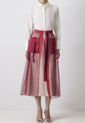 Femme portant une robe blanche à manches longues avec une jupe rouge transparente superposée, dotée de grandes poches et d'une ceinture assortie, associée à des talons blancs.