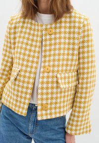 Gul och vit hounds-tooth mönstrad kavaj med kort silhuett, rund halsringning, tre knappar och två framfickor. Avslappnad stil.