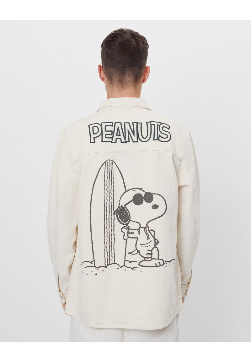 Krämfärgad denimjacka med en stor svart grafik av Snoopy med solglasögon och surfbräda på baksidan. "PEANUTS" är tryckt ovanför.