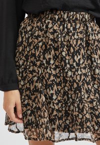 Femme portant une jupe courte avec un imprimé animal abstrait noir, marron et beige et un haut noir à manches longues, montrée de la taille jusqu'à mi-cuisse.