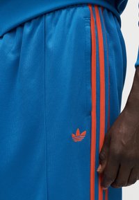 Μπλε αθλητική ενδυμασία με πορτοκαλί ρίγες και κεντημένο λογότυπο. Το υλικό φαίνεται υφή, με ορατές τσέπες στη μέση.